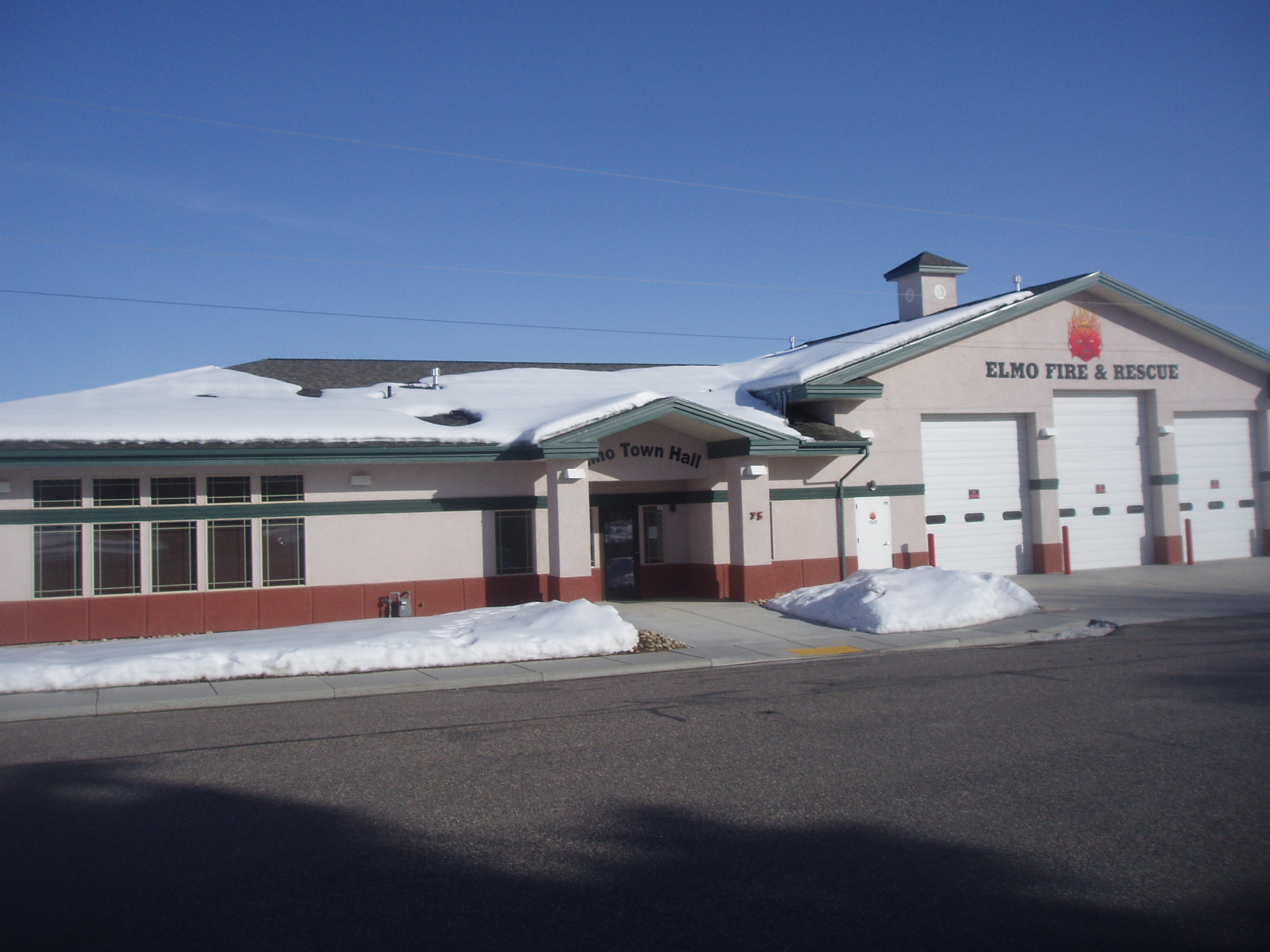 Front of the Elmo Town Hall and Elmo Fire & Rescue building in Elmo, Utah, with snow on the roof, a pedestrian entrance beneath a gable, and three white fire-engine bay doors at right.