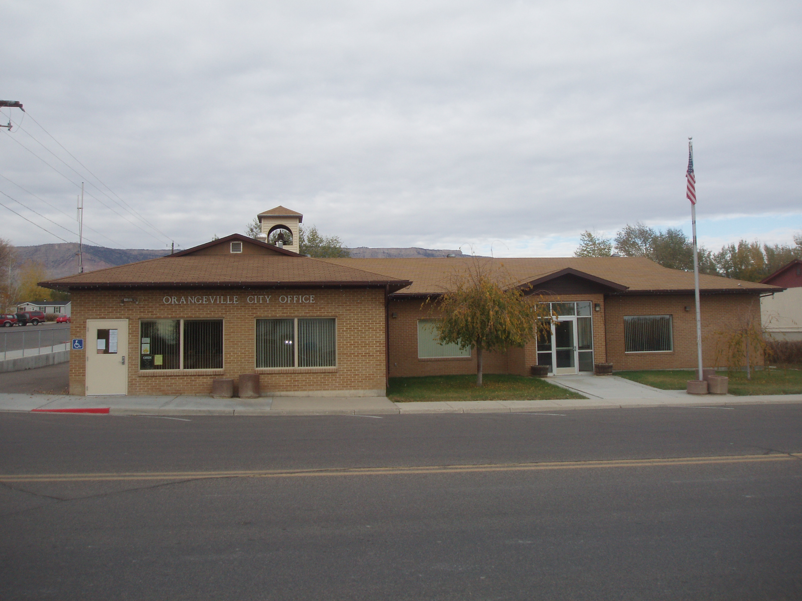 Orangeville City Office, a single-story brick building with a small bell tower, a flagpole at right, and cliffs visible in the distance behind it.