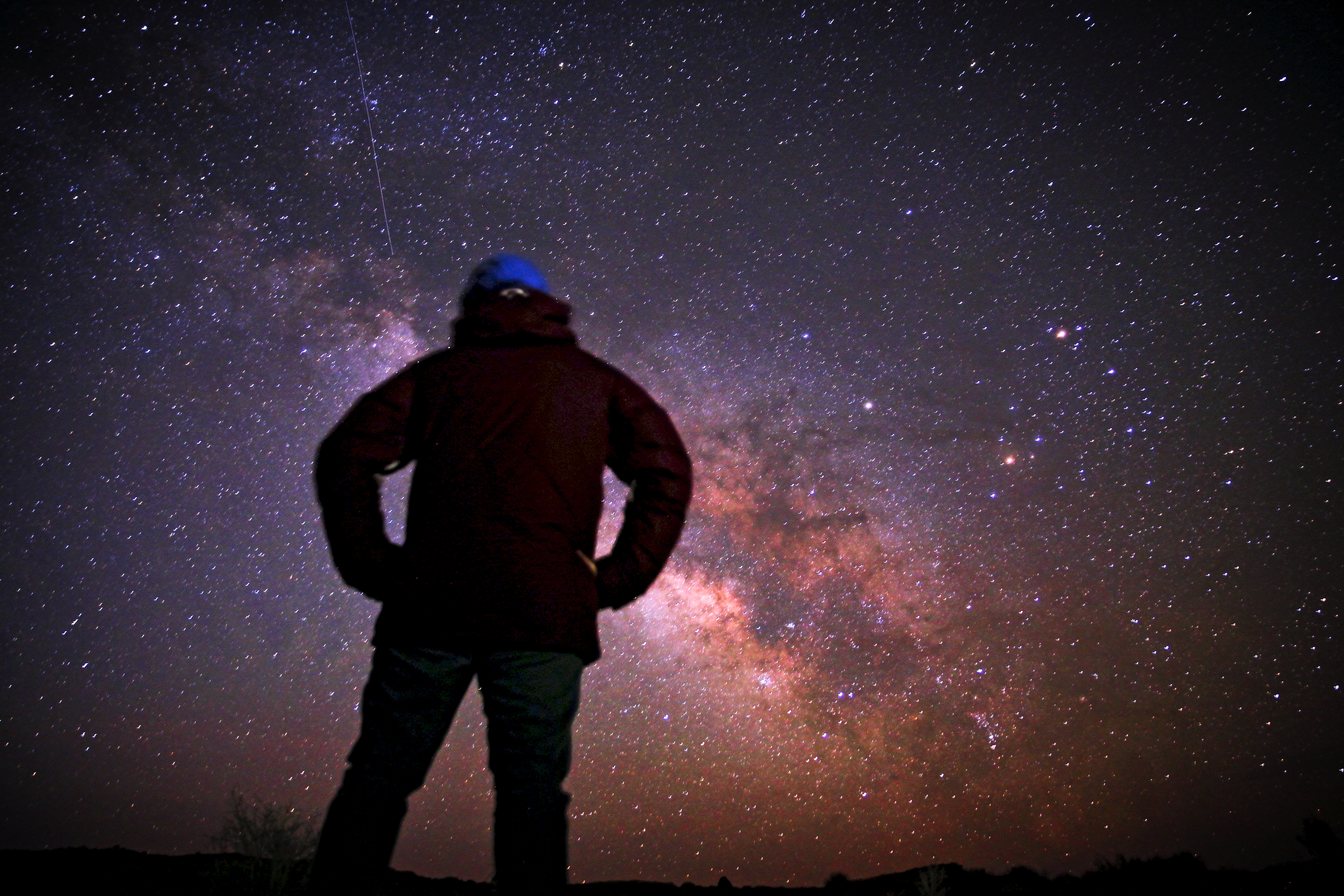 A person in a jacket and cap, seen from behind with hands on hips, looking up at the Milky Way under a dense field of stars, with a thin meteor or satellite trail at upper left.