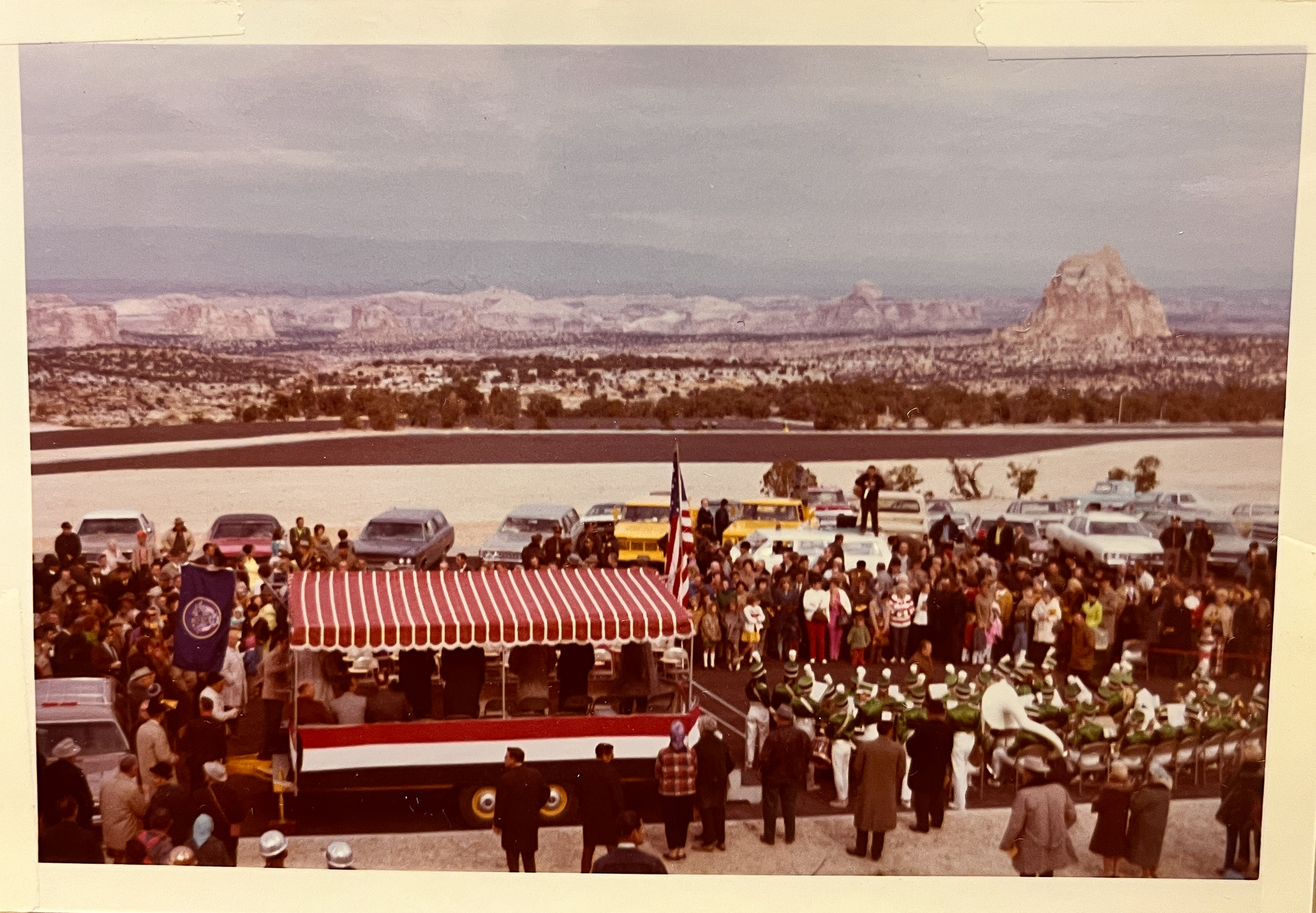 Crowd gathered at the 1970 Interstate 70 dedication at the Ghost Rock rest area: a red-and-white striped speakers' platform with a flag, a green-uniformed band seated at right, parked cars behind, and the San Rafael Swell stretching to a tall rock butte on the horizon.