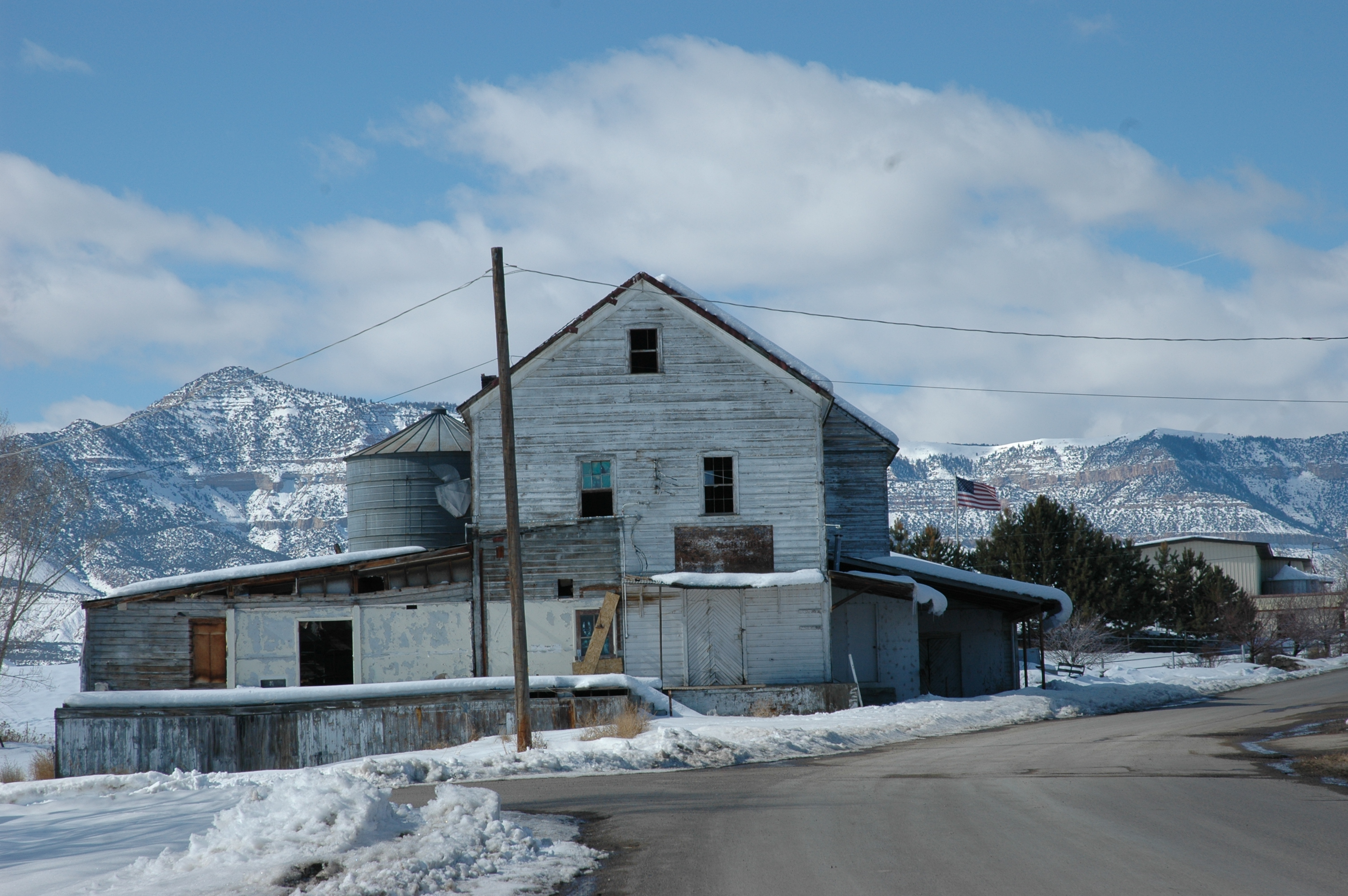 Snow-dusted, weathered white wooden Huntington Roller Mill with a tall gabled center, a metal grain bin at left, and snow-covered mountain cliffs rising behind it, seen from a paved street.