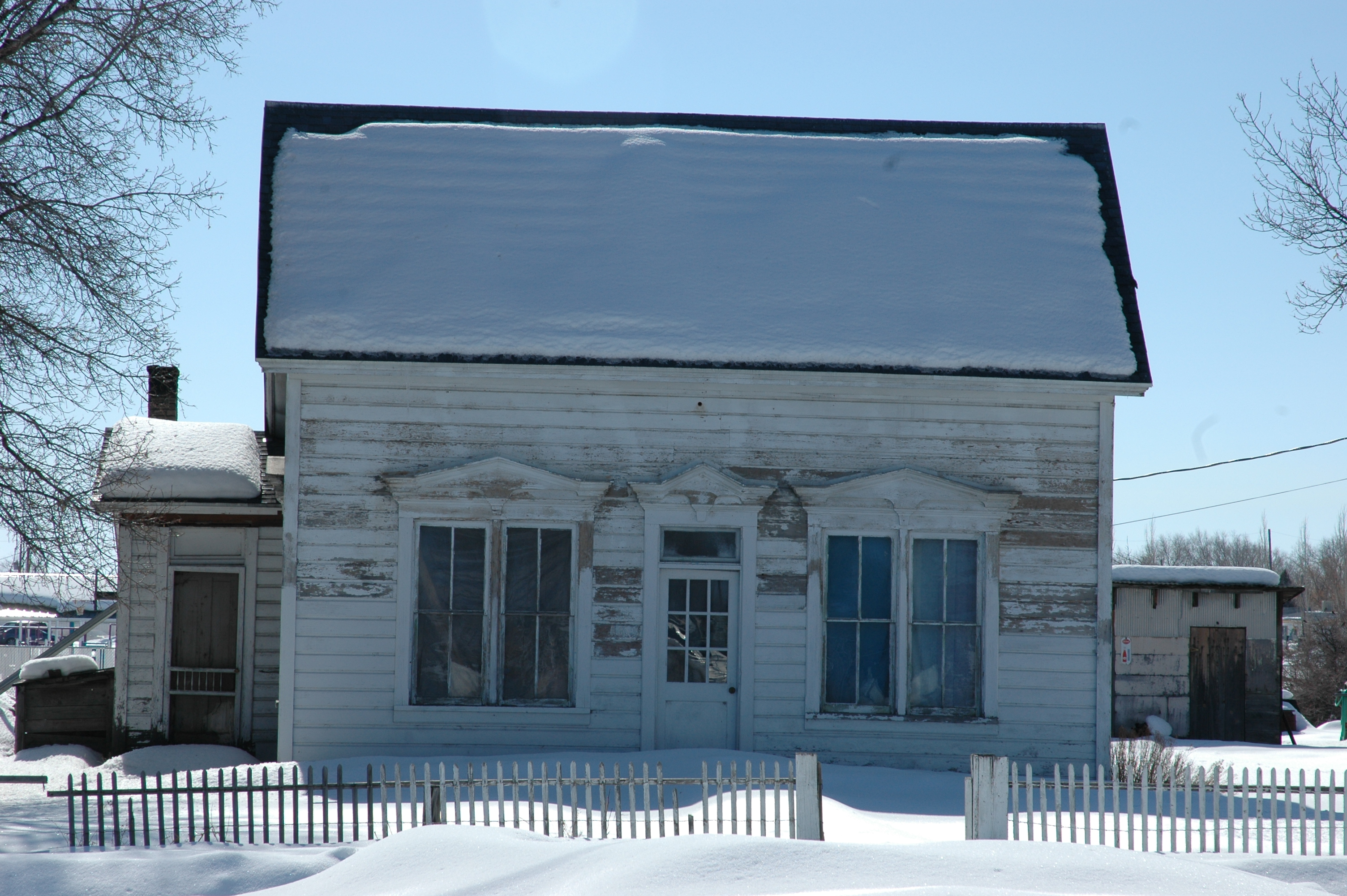 Front of the Leander Lemmon House in winter: a white clapboard house with peeling paint, a snow-covered gable roof, two tall paned windows and a central door under decorative pediments, behind a white picket fence and deep snow.